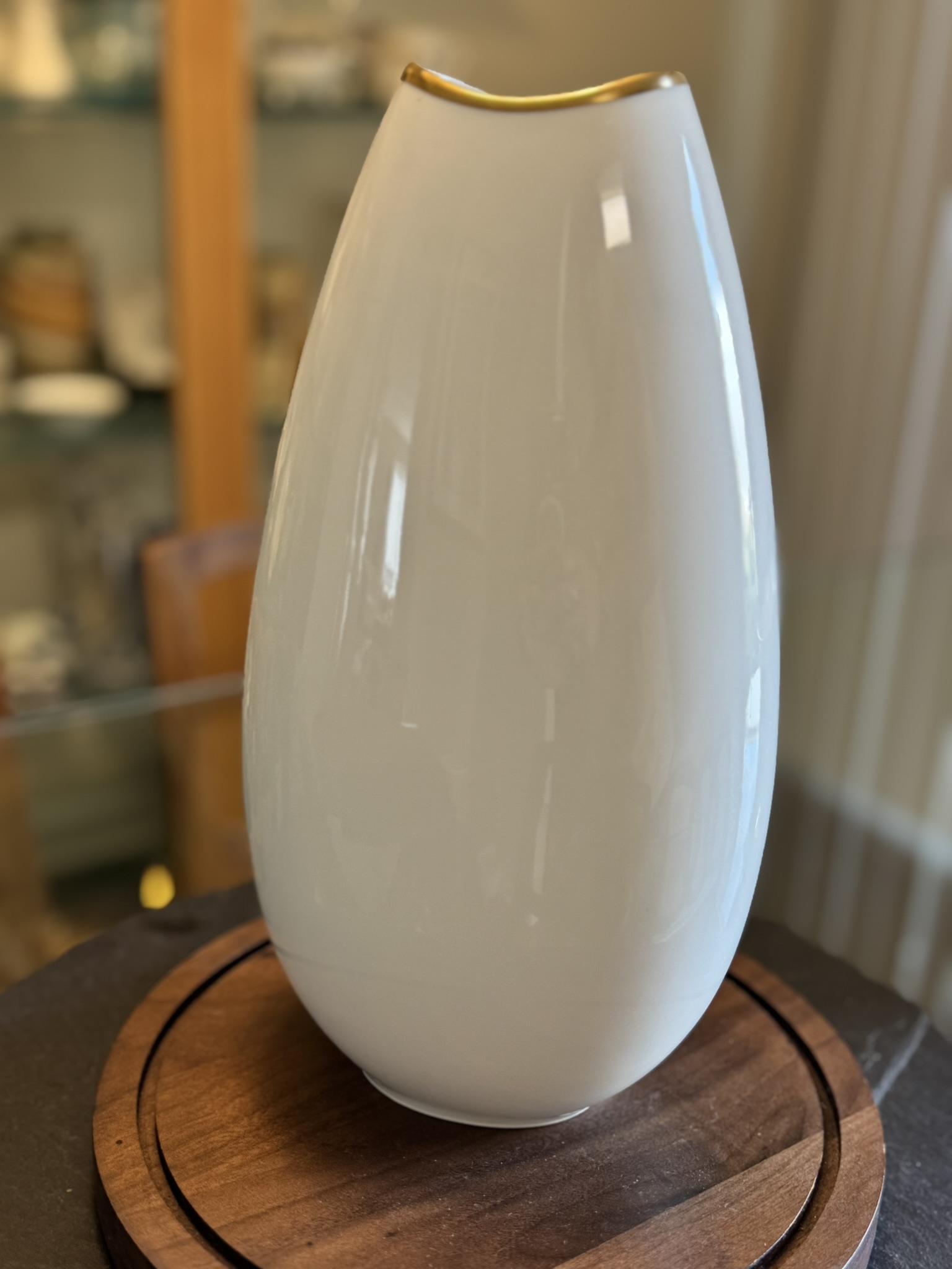 A glossy, white, oval-shaped vase with a narrow golden-rimmed opening sits on a round wooden base. The background shows blurred shelves with assorted pottery and tableware, and soft natural light comes from the right.