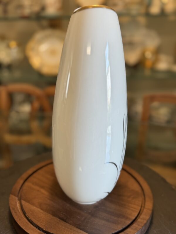A tall, white ceramic vase with a smooth surface and subtle gold rim sits on a round wooden base. The background shows blurred shelves with decorative plates and chairs, giving a warm, vintage atmosphere. Subtle dark lines accent the vases surface.