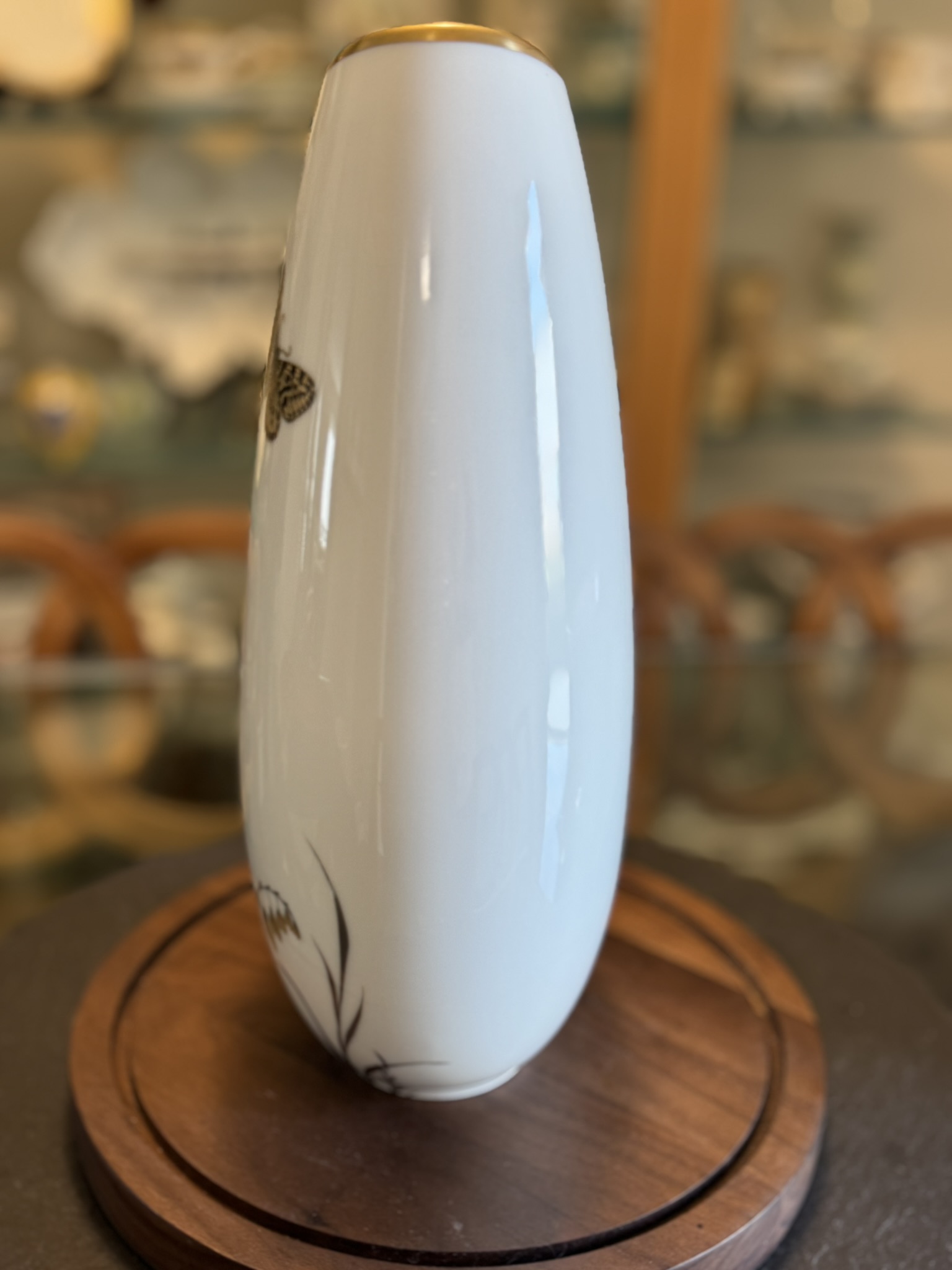 A tall, glossy white vase with a gold rim stands on a round wooden base. The vase features a delicate black floral design and a small butterfly. In the blurred background, ornate shelves hold various ceramics and glassware.