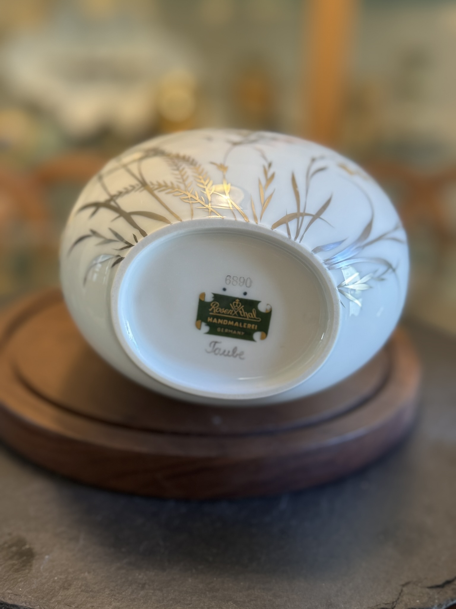 A porcelain bowl rests upside-down on a wooden base, revealing a white bottom with the maker’s mark, gold detailing of wheat and grass, and a green sticker reading “Rosenthal Handmalerei Germany.” The word “Teute” is handwritten on the base.