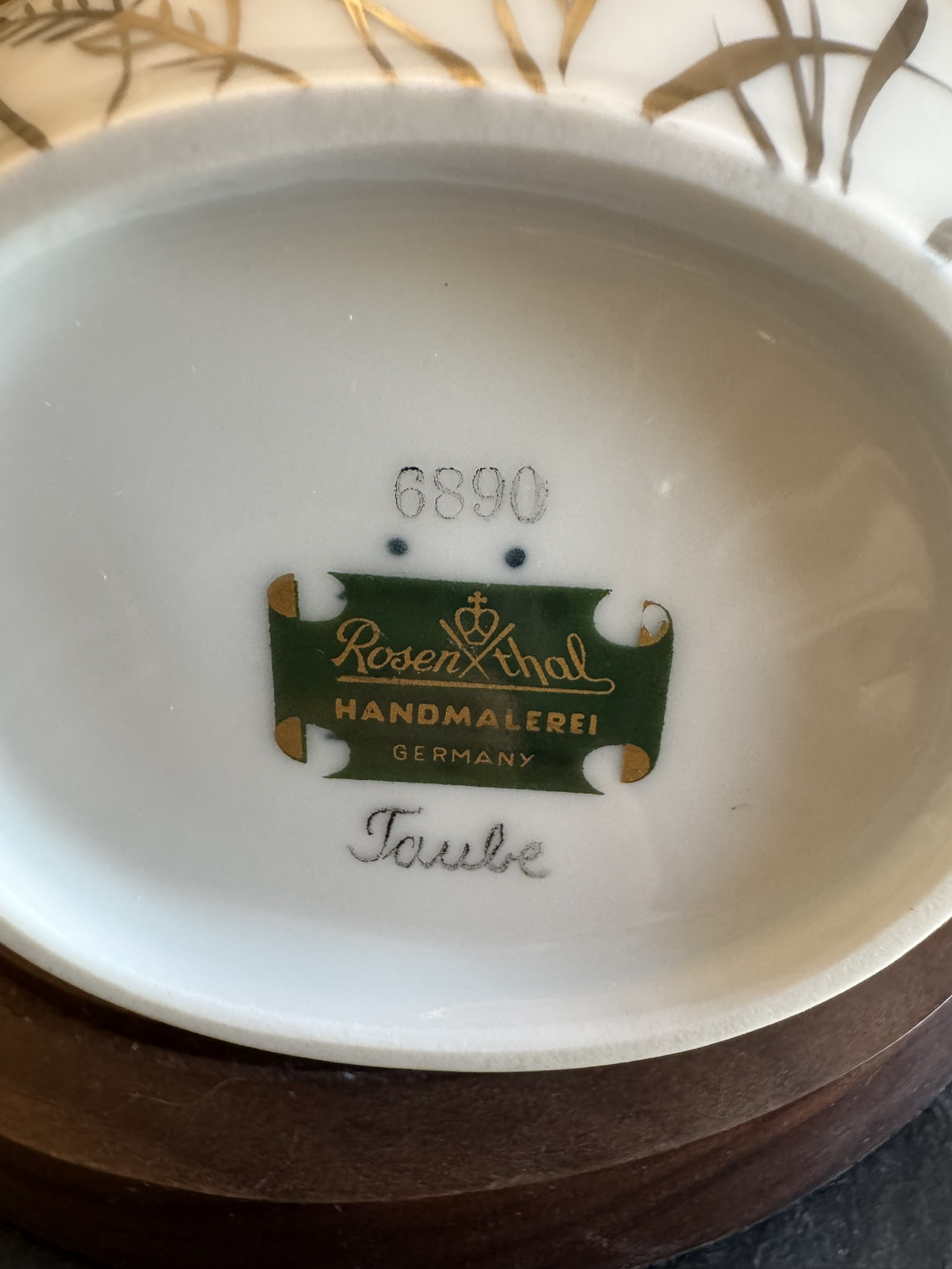Close-up of the bottom of a porcelain item showing a green and gold Rosenthal Handmalerei Germany label, the number 6890 stamped above it, and the word Taube handwritten below on the white glazed surface, with part of a wooden base visible.