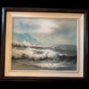 A framed painting depicts crashing ocean waves against rocks under a cloudy sky. In the distance, a white lighthouse stands atop a green cliff. The scene conveys a dramatic, moody seascape with foamy waves and mist. The artist’s signature is visible bottom left.