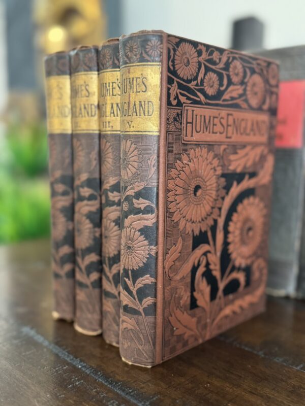 Four ornate, vintage books titled Hume’s England stand upright on a wooden surface. The covers feature embossed floral patterns and golden title labels on the spines. The background is blurred, highlighting the intricate book designs.