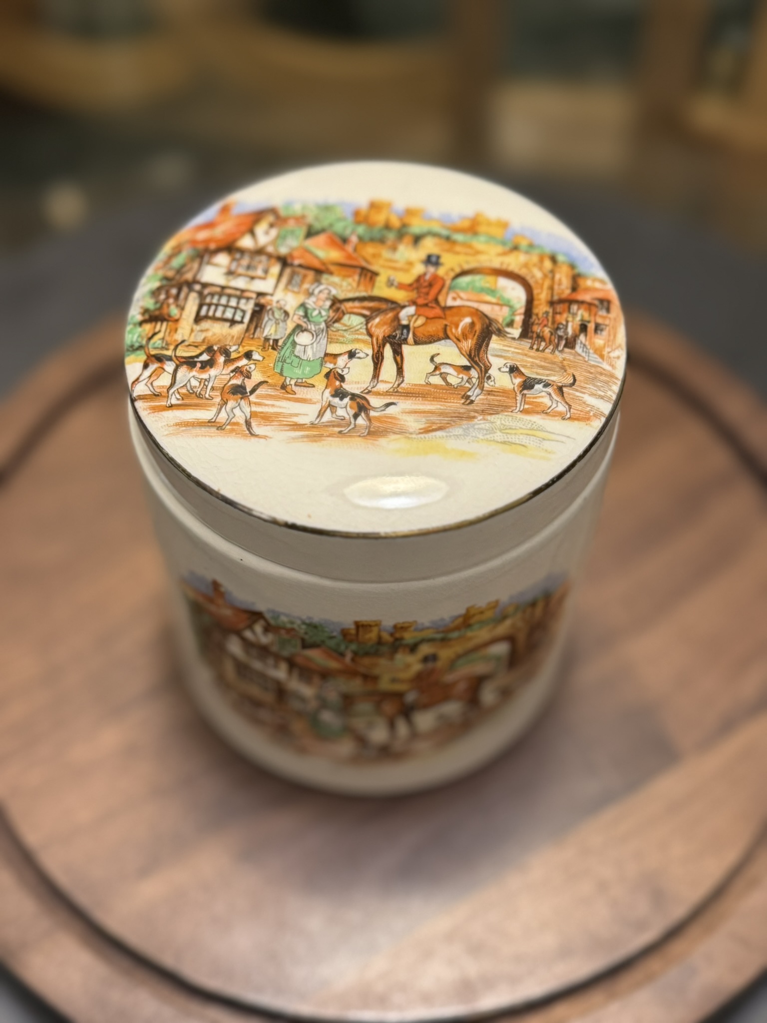A round ceramic container with a lid featuring a colorful painted scene of a village, including people, horses, dogs, and buildings. The container sits on a wooden surface, and the focus is on the detailed lid artwork.