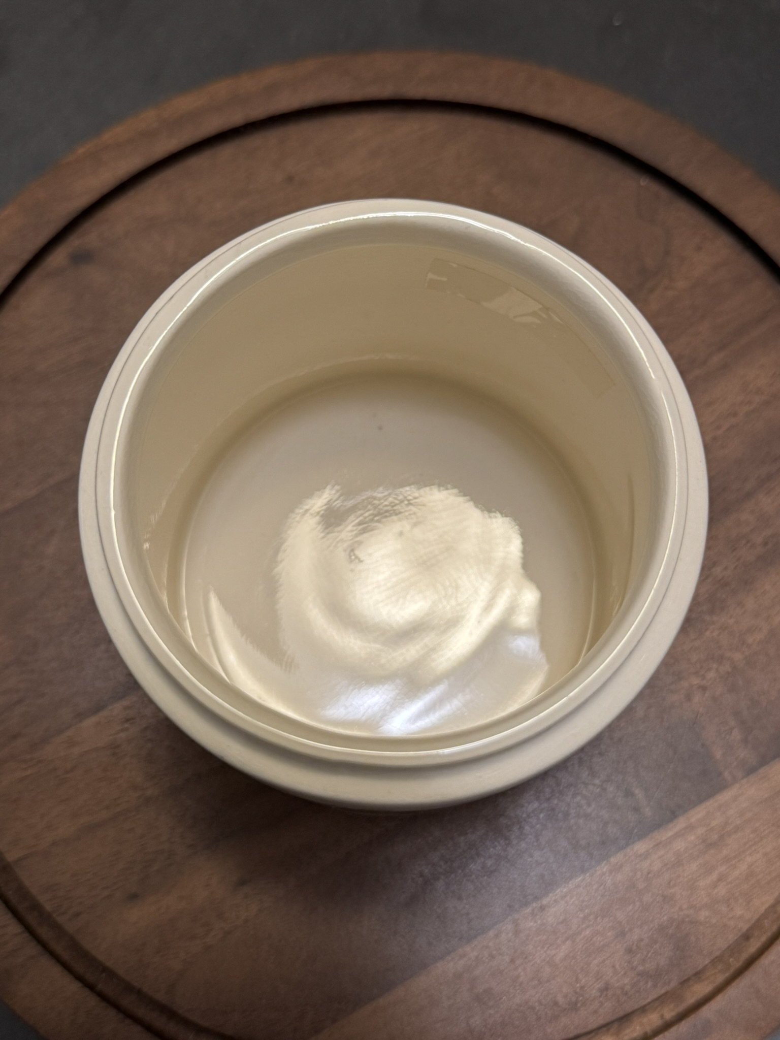 A close-up photo of an open, round white container with a glossy cream or balm inside, placed on a circular wooden surface. The cream appears smooth and untouched, with light reflecting off its surface. The background is a darker wooden table.