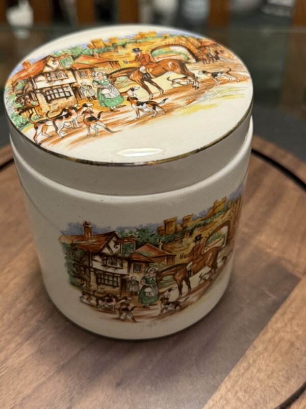 A round ceramic jar with a matching lid, decorated with colorful illustrations of a village scene featuring people, horses, dogs, and cottages. The jar sits on a wooden surface, and the artwork has a vintage, storybook style.