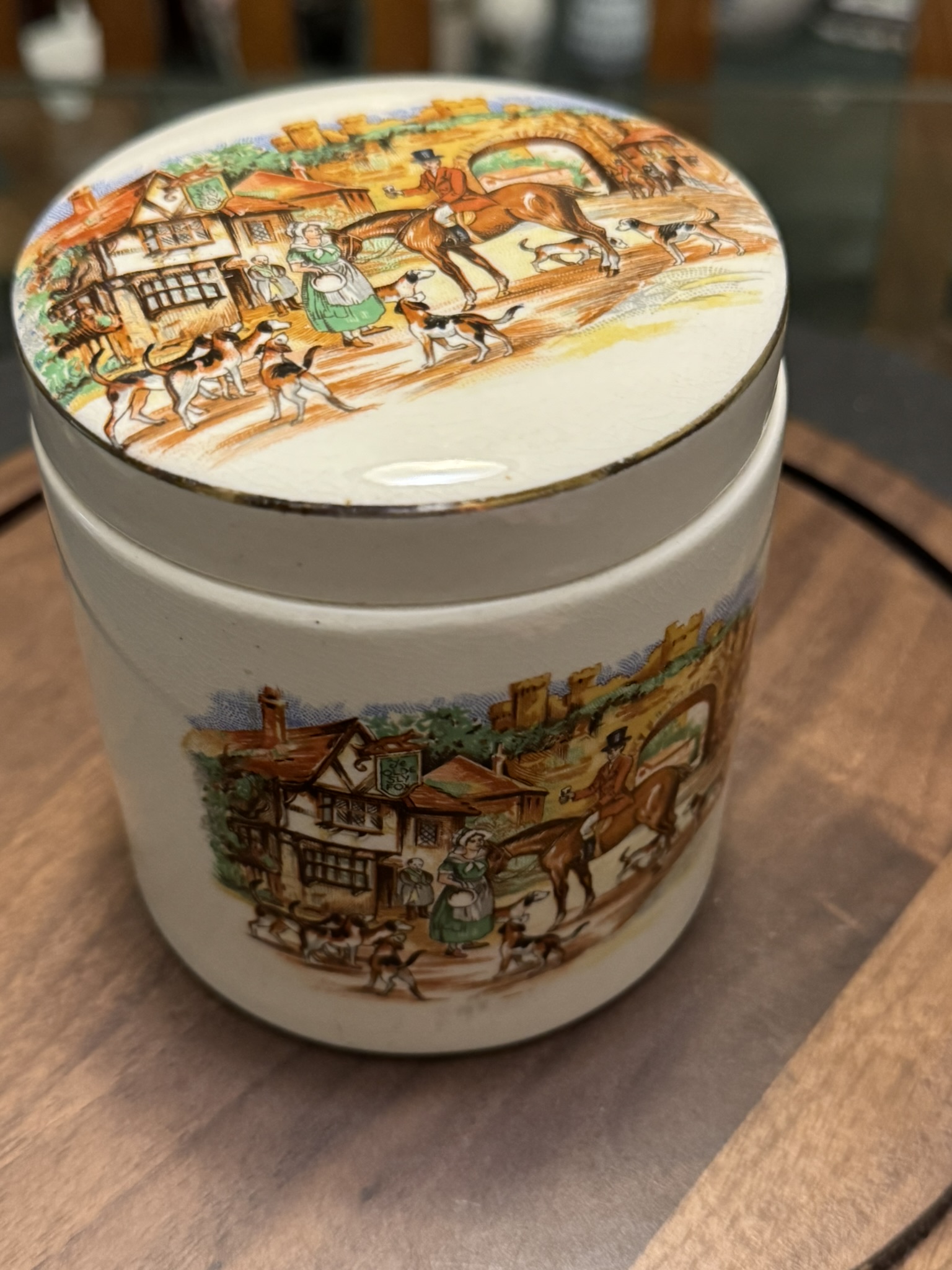 A round ceramic jar with a matching lid, decorated with colorful illustrations of a village scene featuring people, horses, dogs, and cottages. The jar sits on a wooden surface, and the artwork has a vintage, storybook style.
