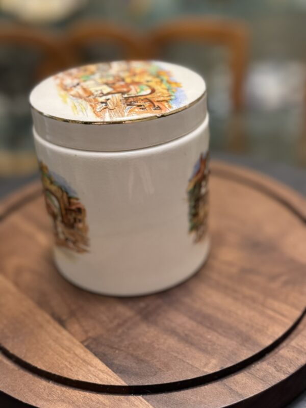 A white ceramic jar with colorful painted scenes on its lid and sides sits on a round wooden surface. The jar appears vintage, with illustrations depicting a rustic village or town. The background is softly blurred, focusing attention on the jar.