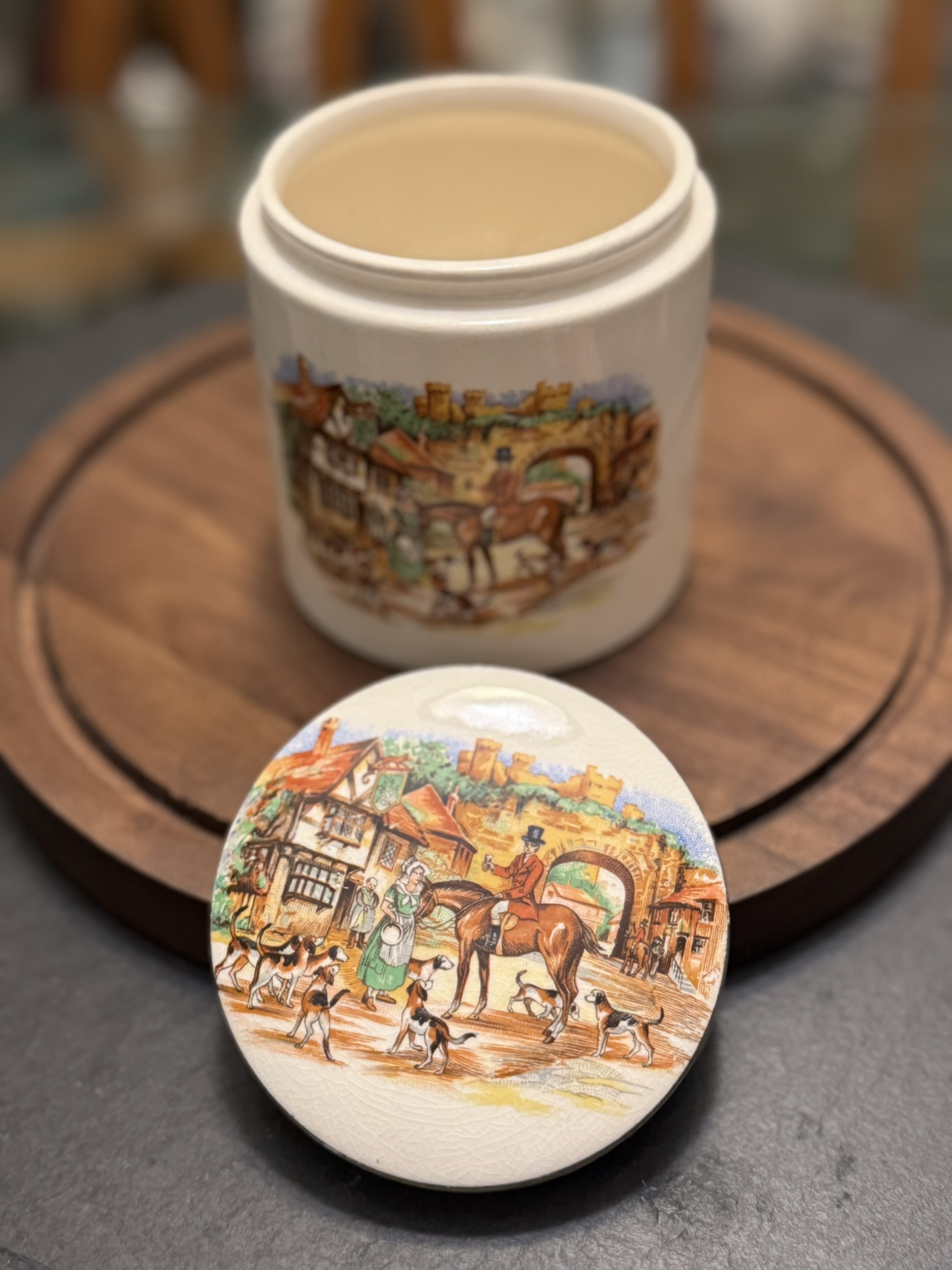 A ceramic jar with a matching lid sits on a wooden surface. Both jar and lid feature colorful illustrations of people, dogs, and horses in a village scene with historic buildings in the background. The lid rests in front, revealing the open jar behind it.