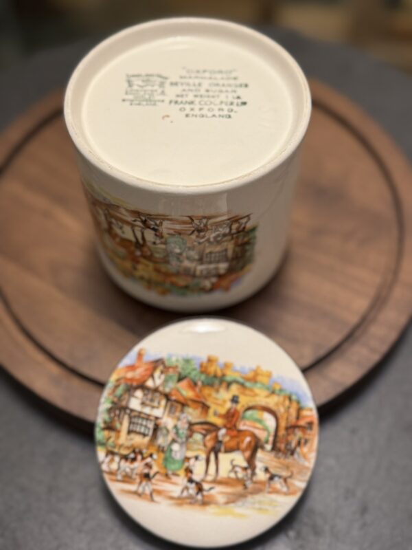 A ceramic jar with a colorful village scene and a lid depicting a horse, people, and dogs in front of cottages. The jar sits on a round wooden tray, and its base shows printed text indicating origin and manufacturer.