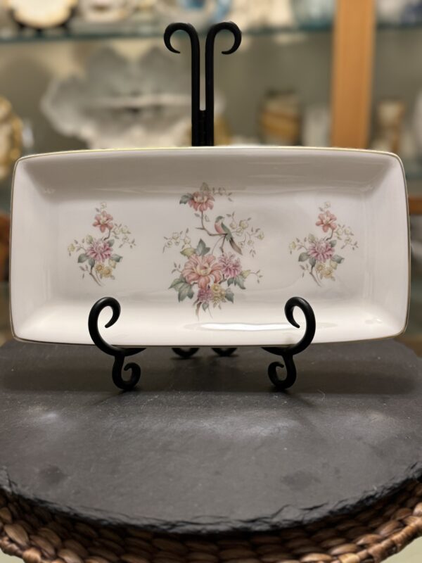 A rectangular white porcelain plate with delicate pink and green floral designs sits upright on a black metal stand. The background is softly blurred, showing shelves with various ceramics and decorative items.