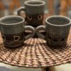 Three textured ceramic mugs with gray upper halves and brown lower halves, featuring gold accents, sit on a round woven mat. The background is softly blurred, showing wooden chairs and a table.
