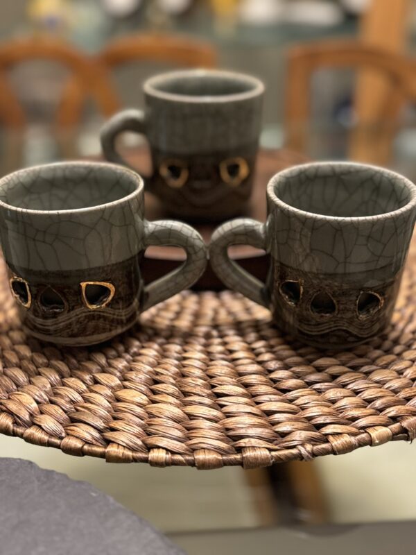 Three textured ceramic mugs with gray upper halves and brown lower halves, featuring gold accents, sit on a round woven mat. The background is softly blurred, showing wooden chairs and a table.