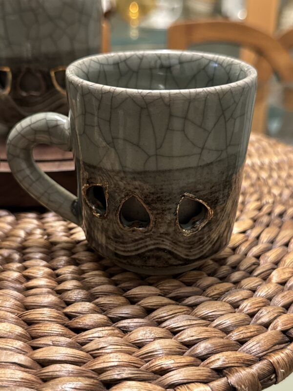 A ceramic mug with a crackled gray top and textured dark brown base sits on a woven placemat. The base has three teardrop-shaped cutouts edged in gold, and a curved handle is visible on the left side. Another mug is partially visible in the background.