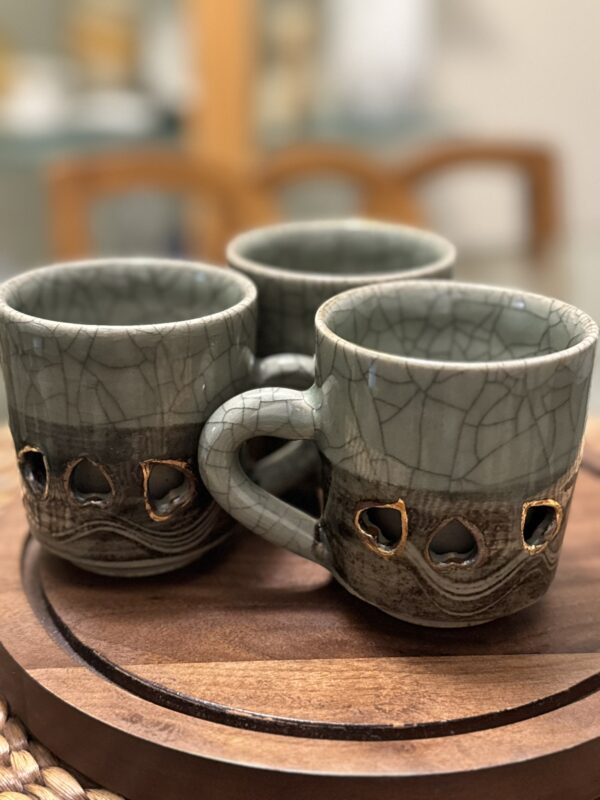 Three gray ceramic mugs with a cracked, textured glaze and heart-shaped cutouts are arranged in a triangle formation on a round wooden tray. The background is softly blurred, highlighting the mugs artistic design.