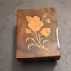 A small, rectangular, dark brown wooden box sits on a dark surface. The box lid features an inlaid floral design with two yellow-orange flowers and green stems and leaves, creating a vintage, decorative look. The wood has a glossy finish.
