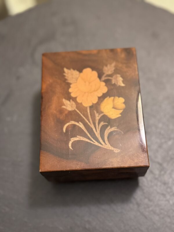 A small, rectangular, dark brown wooden box sits on a dark surface. The box lid features an inlaid floral design with two yellow-orange flowers and green stems and leaves, creating a vintage, decorative look. The wood has a glossy finish.