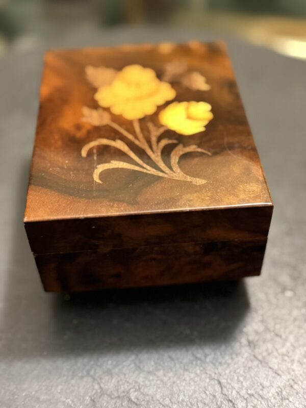 A small, rectangular wooden box with a glossy finish. The lid features an inlaid floral design with yellow and orange flowers and greenish stems, set against a rich, brown wood grain background. The box rests on a dark, slightly textured surface.