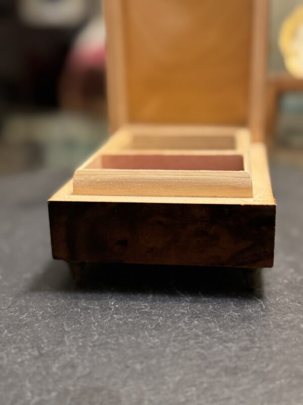 A close-up of an open wooden box with a hinged lid, viewed from the front. The box has square compartments inside and sits on a dark, textured surface; the background is softly blurred.