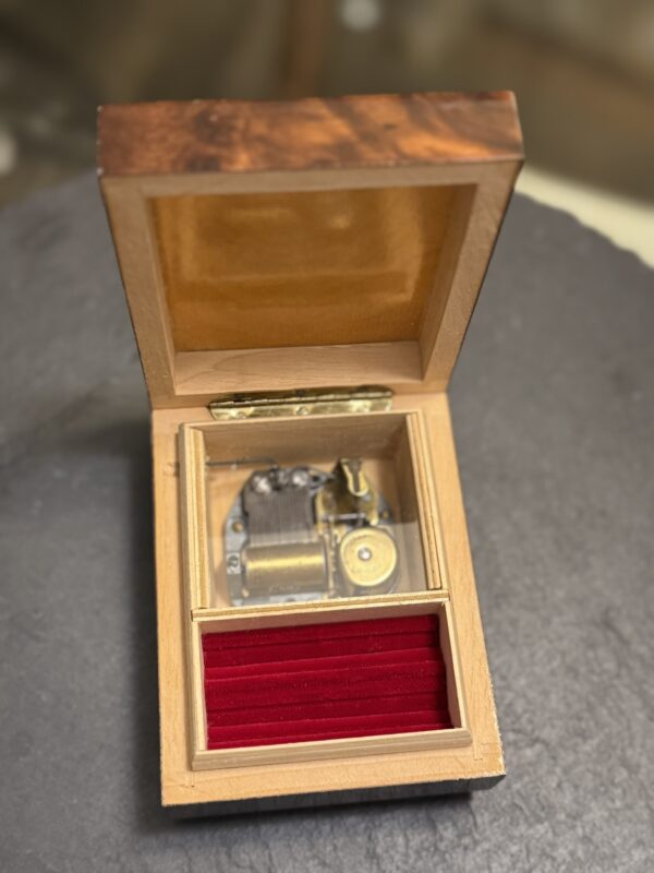 A wooden music box with its lid open sits on a dark surface. Inside, the metal music mechanism is visible on the left, and a red velvet-lined compartment is on the right. The box’s interior is smooth and polished, with yellow felt on the lid’s inner side.