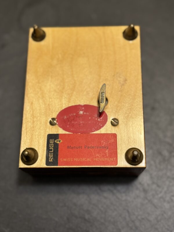 A wooden box sits upside down showing four brass feet, a metal winding key, and a red label reading “REUGE Menuet Paderewski SWISS MUSICAL MOVEMENT.” The wood is pale with visible grain, on a dark surface.