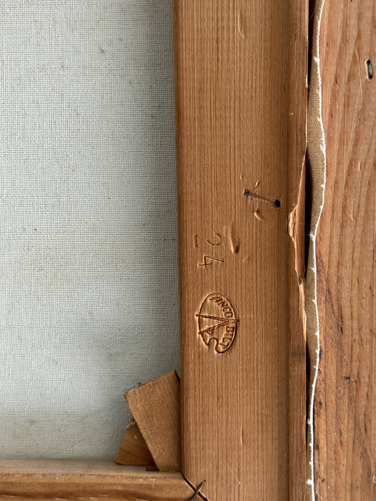 Close-up of the back corner of a wooden picture frame with canvas, showing a torn, folded edge, a visible nail, and a round stamp with text and numbers 24 carved into the wood. The texture of the wood and canvas is clearly visible.