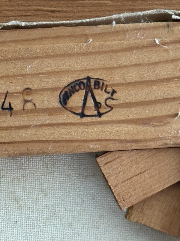 Close-up of a wooden stretcher bar with a black ink stamp reading “AMPCO BILT” in an oval shape, with a stylized “A” at the center. Part of a canvas and some overlapping wood pieces are visible in the image.