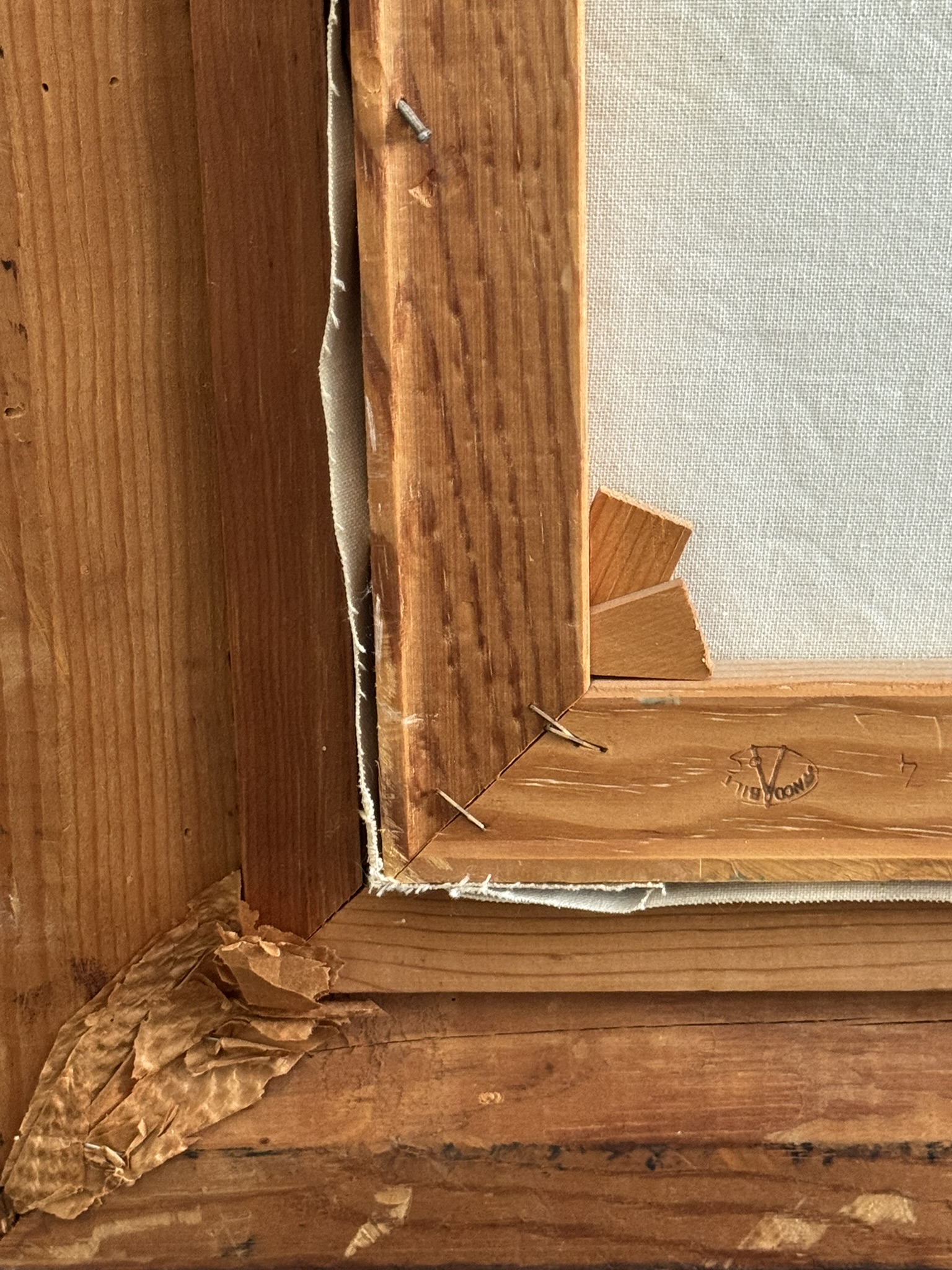Close-up of the back corner of a wooden picture frame, showing staples securing a canvas. The wood has a rough texture, some chipped sections, and a paper tape corner. A faint stamp reading “FIR WOOD” is visible on the wood.
