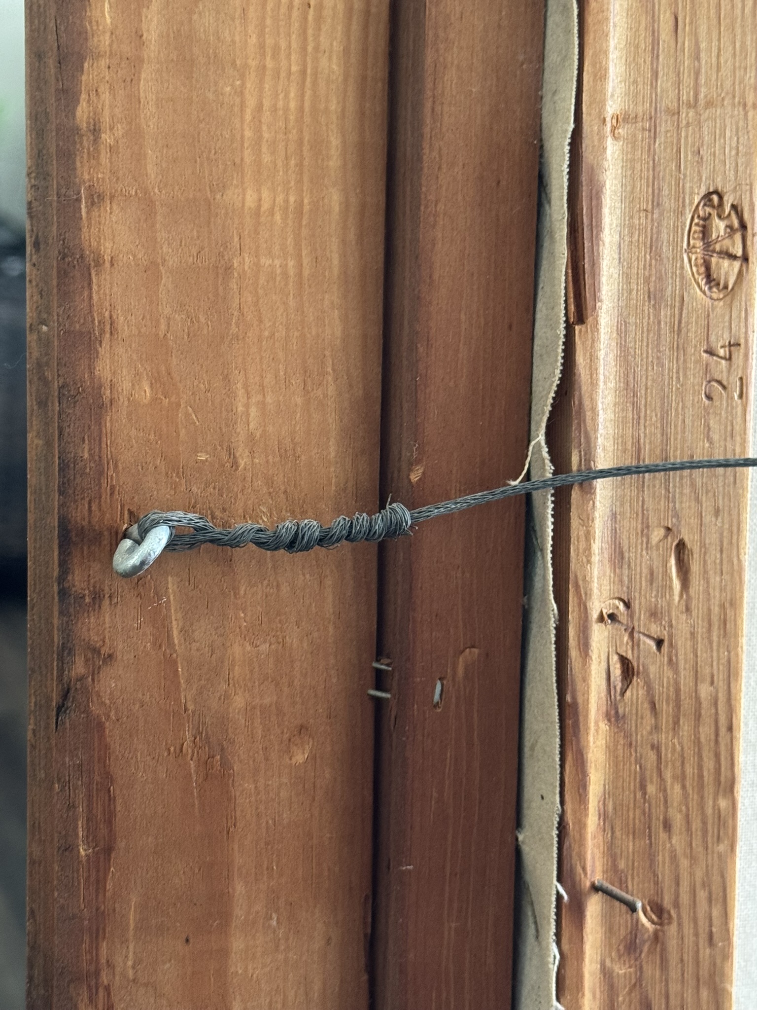A close-up of a wire twisted around a metal nail in a wooden frame, likely for hanging the frame. The wood is medium brown with visible grain, some wear, and a stamped marking reading 24 on the right edge. Cardboard backing is partially visible.