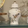 A wooden and glass display shelf filled with decorative porcelain items, including floral teacups, plates, a teapot, a white elephant figurine, vases in various shapes and colors, a sea-themed plate, and a gold-trimmed bowl on a stand.