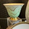 A decorative porcelain bowl with a pale green and gold rim, featuring delicate gold leaf and stem patterns. The bowl sits atop a clear cube stand, with part of a white plate with a gold border visible in the foreground.