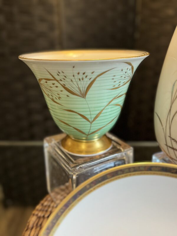 A decorative porcelain bowl with a pale green and gold rim, featuring delicate gold leaf and stem patterns. The bowl sits atop a clear cube stand, with part of a white plate with a gold border visible in the foreground.
