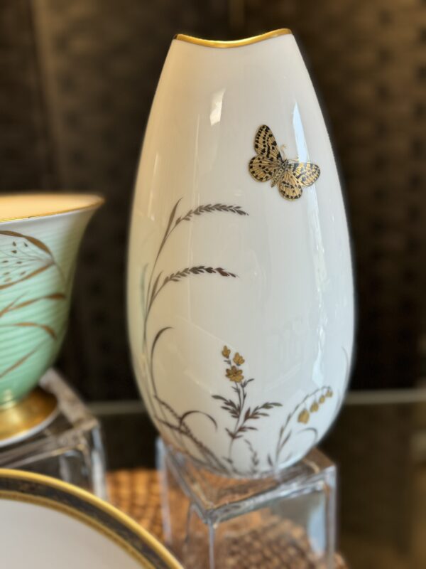 A tall, white porcelain vase with gold trim features a delicate gold and black butterfly. Brown and gold botanical designs, including grasses and small flowers, wrap around the vase. The background includes a mint-green bowl and part of a white plate.