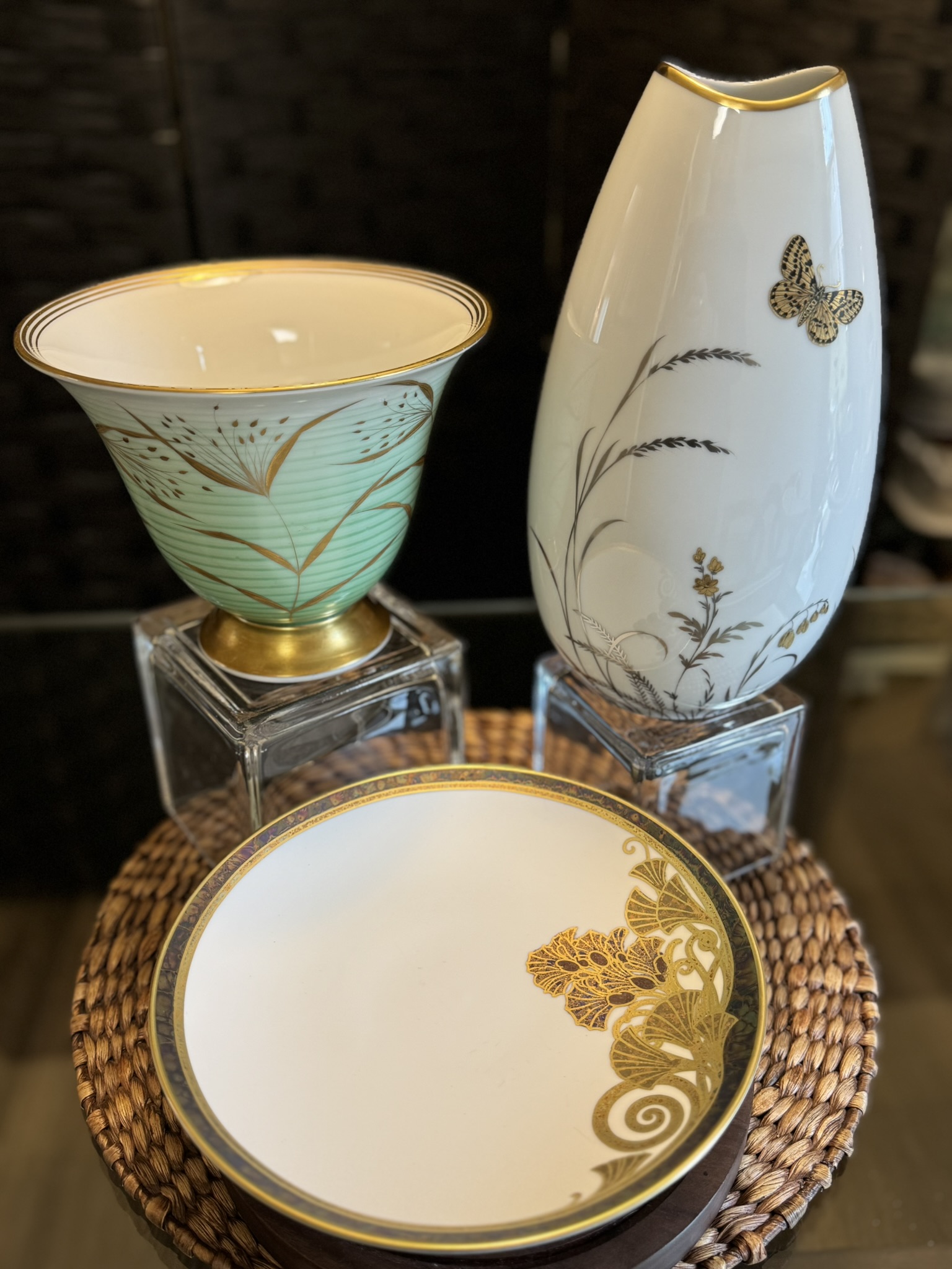 Three elegant ceramics are displayed on a woven mat: a green and gold chalice-shaped vase, a tall white vase with floral and butterfly designs, and a round bowl with a gold floral pattern, each elevated on clear square stands.