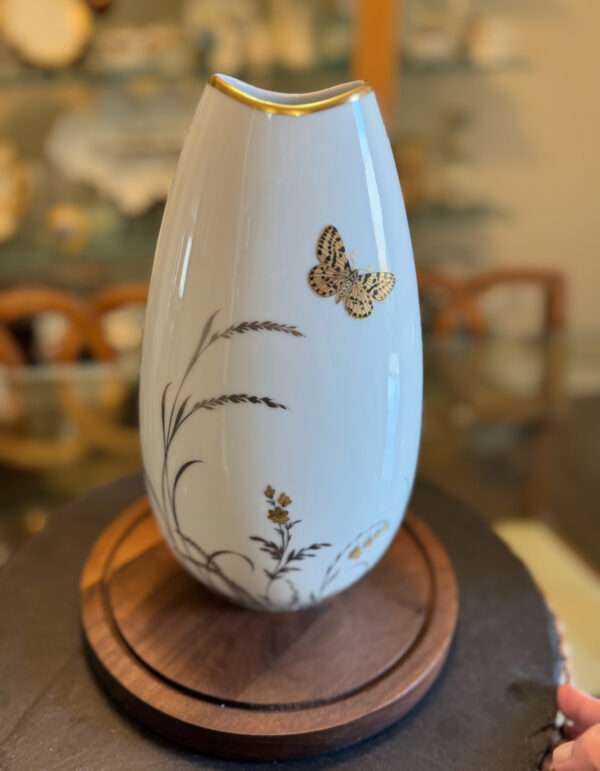 A white, oval-shaped porcelain vase with a gold rim features delicate black and gold botanical designs and a gold butterfly decoration. The vase sits on a round wooden base, with a softly blurred background of shelves and chairs.