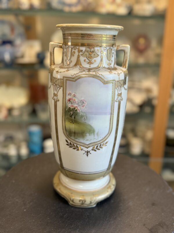 A tall, ornate porcelain vase with gold accents and handles, featuring a painted landscape of pink flowers, green grass, and a distant body of water. The vase stands on a dark surface, with a blurred background of shelves displaying collectibles.
