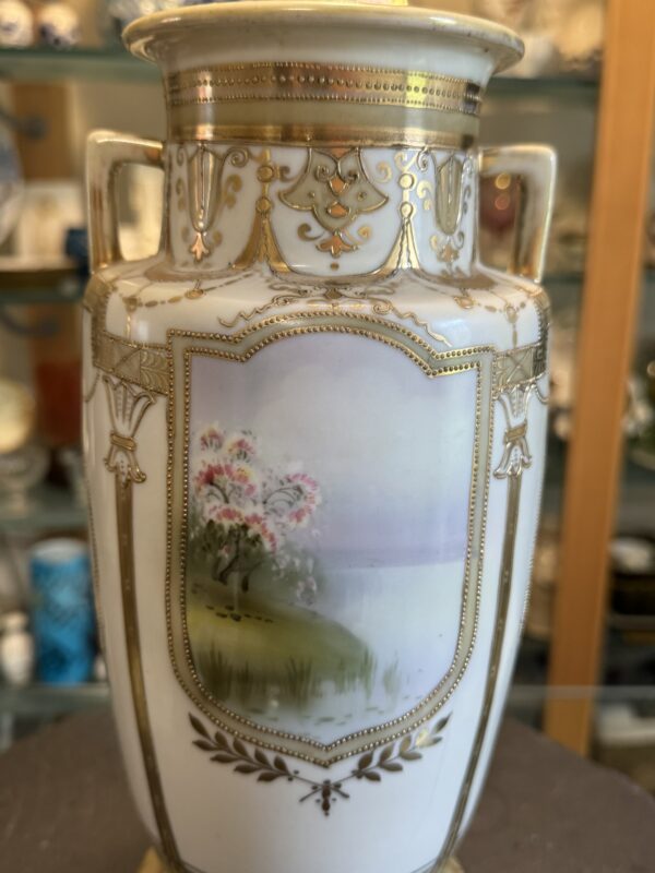 A decorative porcelain vase with gold accents and handles features a painted scene of flowering trees on a grassy bank beside a calm lake, under a soft pastel sky. Ornate gold patterns and trims embellish the white surface around the painting.