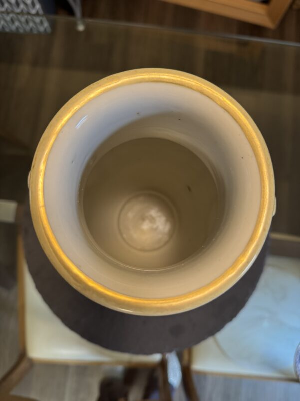 A top-down view of an empty ceramic vase with a light brown rim and a beige interior. The vase sits on a glass table, with part of a chair and wood flooring visible in the background. The lighting suggests an indoor setting.