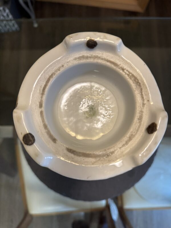 Close-up of the bottom of a ceramic object, possibly a vase or lamp base. The base is white with three dark felt pads spaced around the rim and some dirt or wear marks. The center shows a recessed circular area with a faint green stamp or residue.