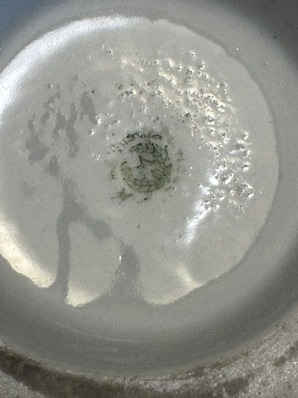 A close-up of the bottom of a shallow gray or off-white pan with a small amount of oil and scattered bits of finely chopped green herbs or spices sizzling in the center. Light reflections and oil bubbles are visible on the pan’s surface.