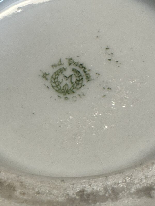 A close-up of the underside of a ceramic plate shows a faded green maker’s mark. The mark includes a laurel wreath encircling a bird with outstretched wings, possibly an eagle. Some parts of the mark are worn and speckled. The plate is off-white.