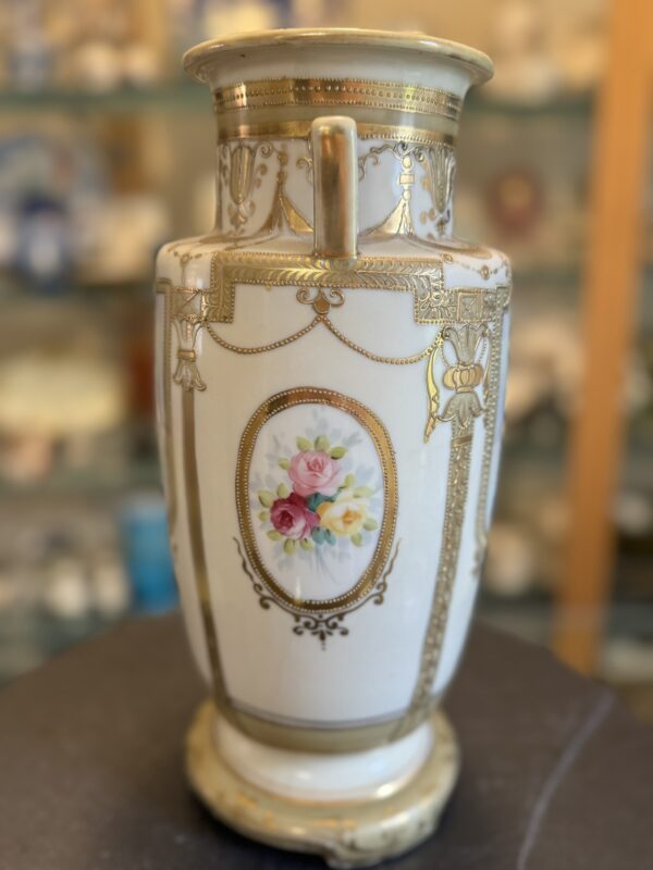 A tall, ornate porcelain vase with gold detailing, vertical handles, and an oval floral motif featuring pink and yellow roses with green leaves. The vase has intricate gold patterns and stands on a round base, displayed indoors against a blurred background.