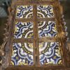 A wooden serving tray with intricate carved edges holds colorful ceramic tiles. The tiles show a symmetrical blue, yellow, and white mandala-like pattern, divided by wooden bars into six sections. The tray rests on a reflective surface.