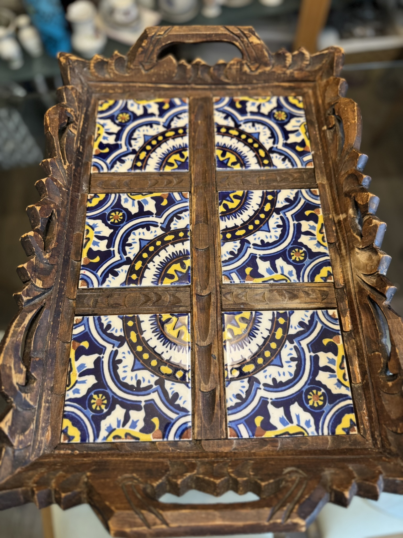A wooden serving tray with intricate carved edges holds colorful ceramic tiles. The tiles show a symmetrical blue, yellow, and white mandala-like pattern, divided by wooden bars into six sections. The tray rests on a reflective surface.