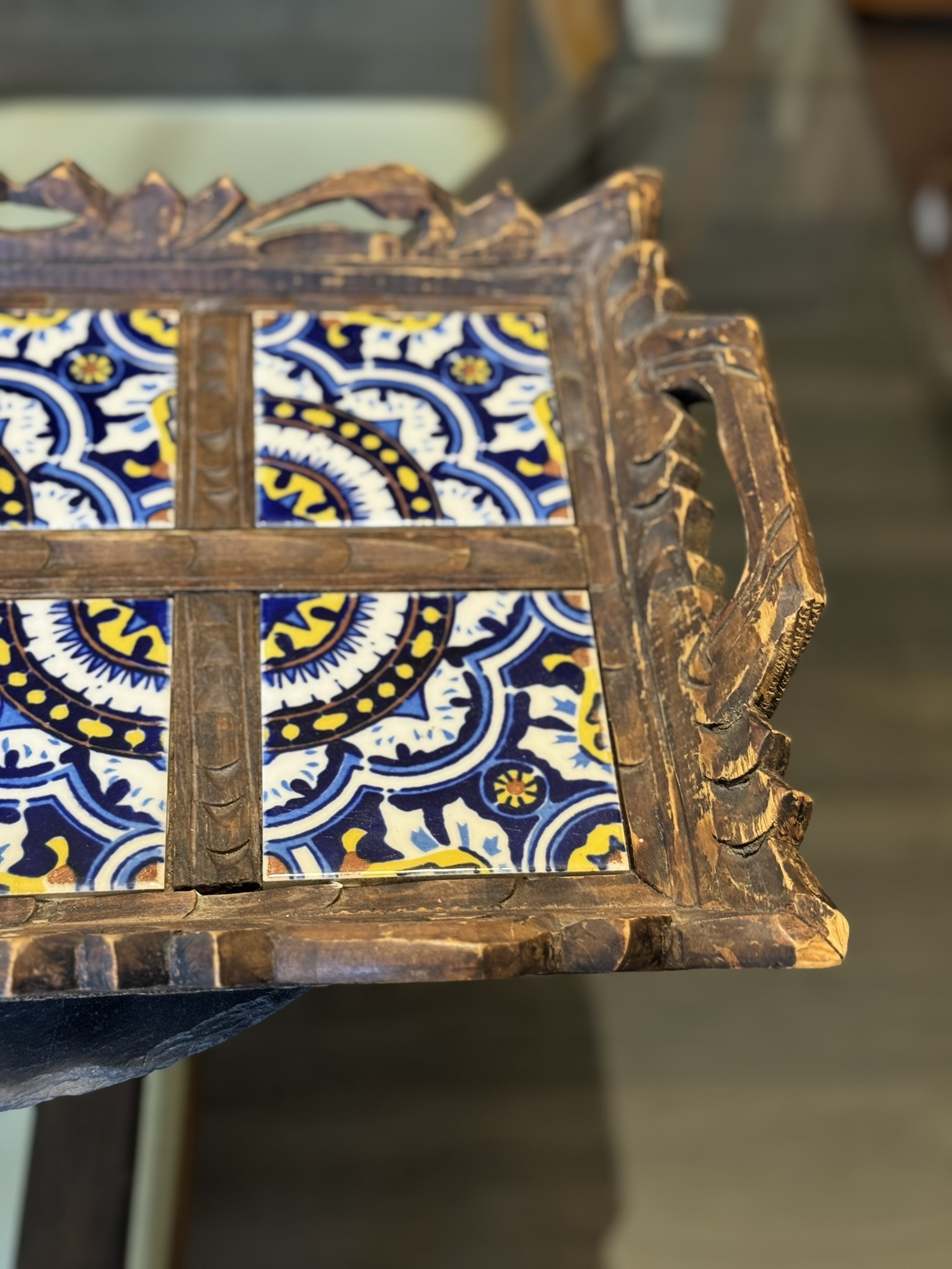 A close-up of a rustic wooden tray with ornate carved handles and edges, featuring colorful ceramic tiles. The tiles display an intricate blue, white, and yellow floral and geometric pattern. The tray rests on a dark surface in a warmly lit room.