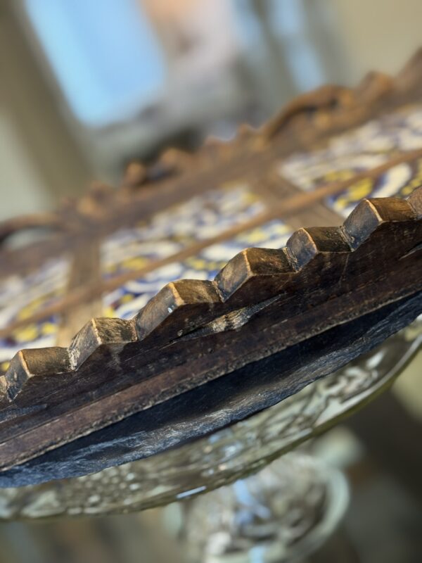 A close-up of a round wooden tray with scalloped edges, featuring colorful, intricate tile inlay. The focus is on the trays edge, highlighting the wood grain and craftsmanship, while the background is softly blurred, suggesting an indoor setting.
