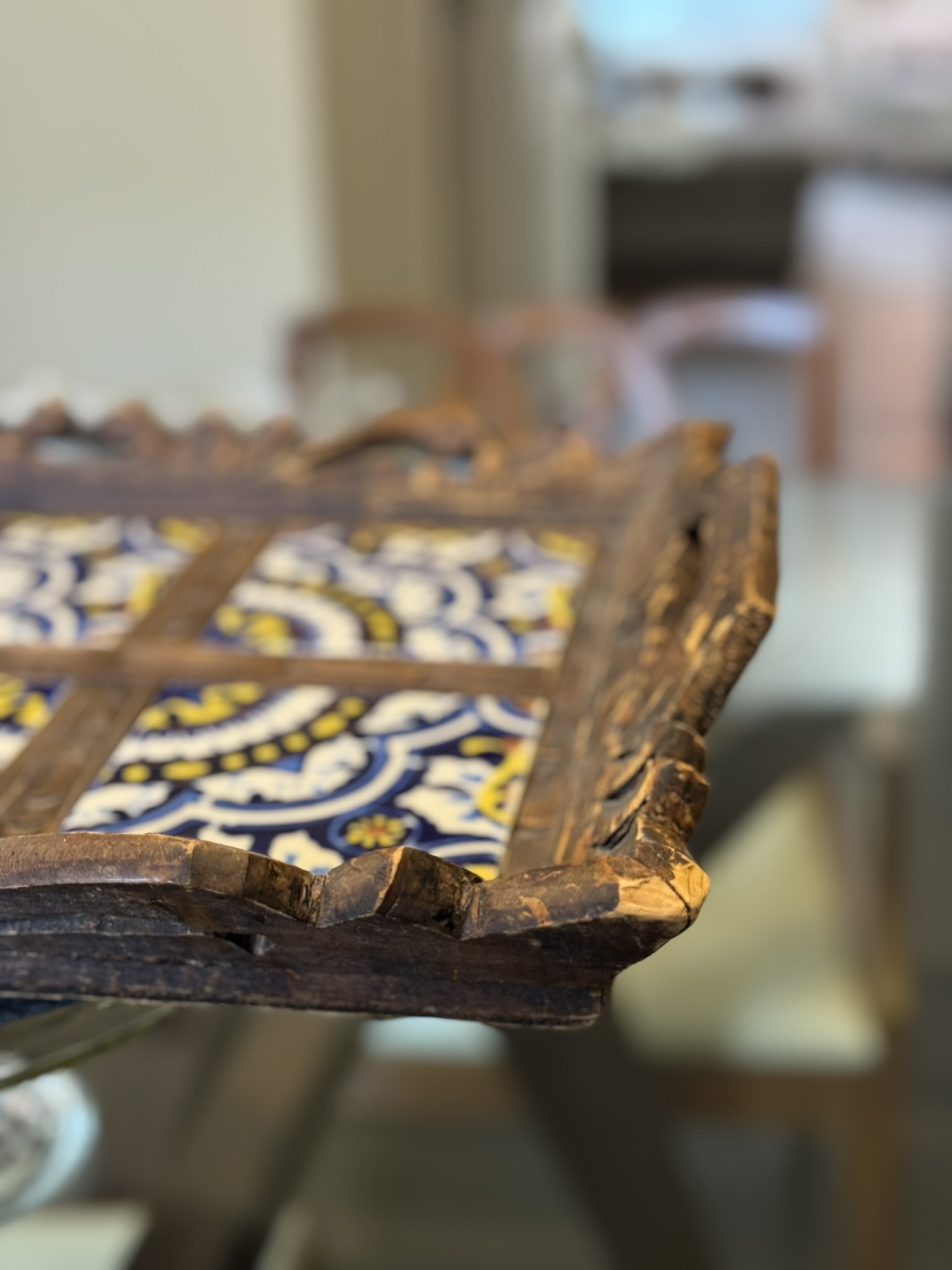A close-up of a rustic wooden tray with colorful blue, yellow, and white patterned tiles. The tray has a carved, weathered edge. The background is softly blurred, showing a table and chairs, giving a cozy indoor setting.