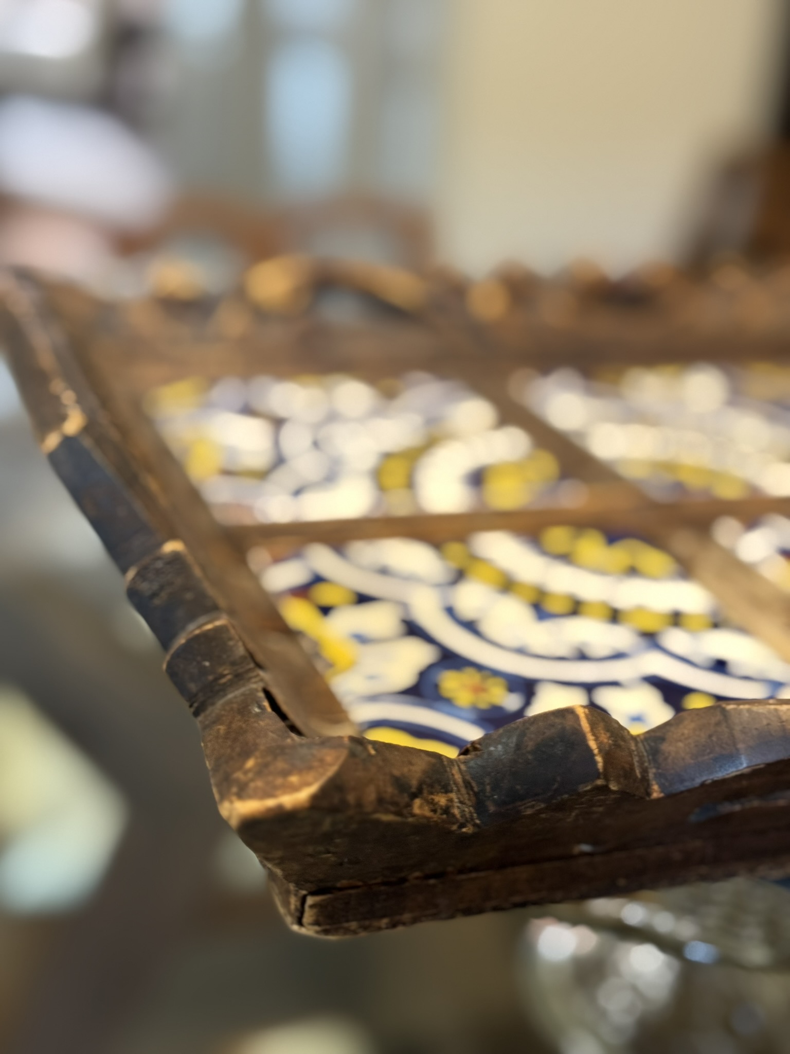 A close-up of a rustic wooden tray with decorative painted tiles. The tiles feature intricate blue, white, and yellow patterns. The wooden edges of the tray appear worn and textured, suggesting age and frequent use. The background is blurred.