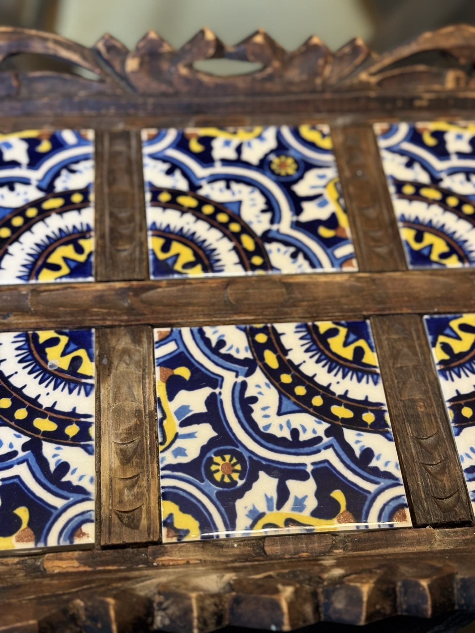 A close-up of a decorative wooden tray features three blue, yellow, and white patterned ceramic tiles with intricate, symmetrical designs, framed by dark brown carved wood slats. The tray edges have ornate carvings.