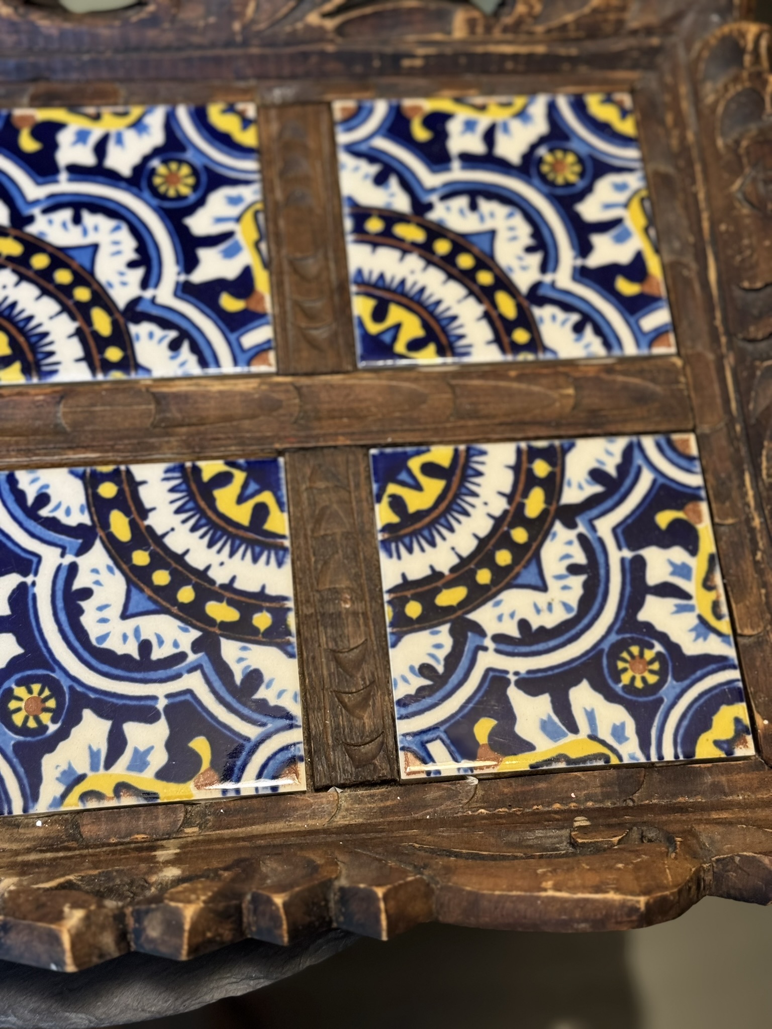 A close-up of a wooden tray featuring a decorative pattern of blue, yellow, and white tiles arranged in four sections, separated by rustic wooden dividers. The intricate design includes geometric and floral motifs, adding a vibrant, handcrafted look to the tray.